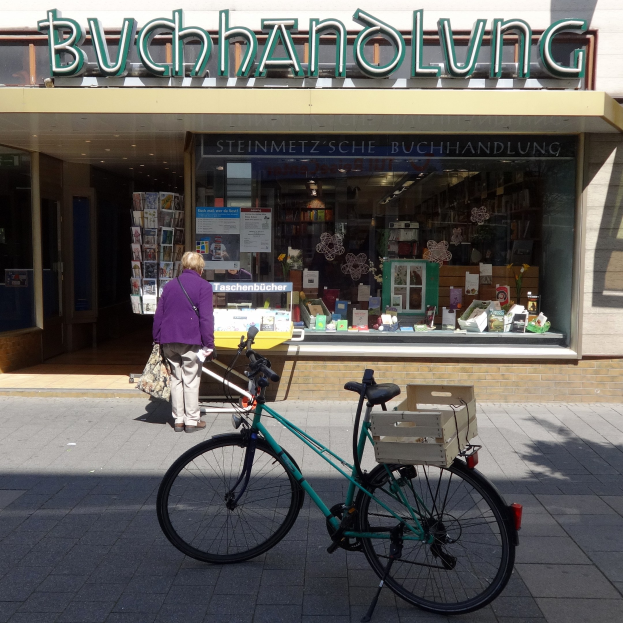 Eine Frau steht neben einem Fahrrad, das vor einem Geschäft geparkt ist. Im Vordergrund ist ein Fahrradrad zu sehen und durch die Glasscheibe des Geschäfts sind Bücher und andere Gegenstände zu erkennen.