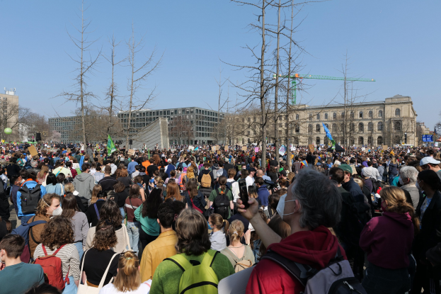 Eine große Menge von Menschen, die an einer Klimaprotestaktion vor einem Gebäude mit Bäumen und einem klaren Himmel teilnehmen, viele halten Schilder und tragen Taschen.