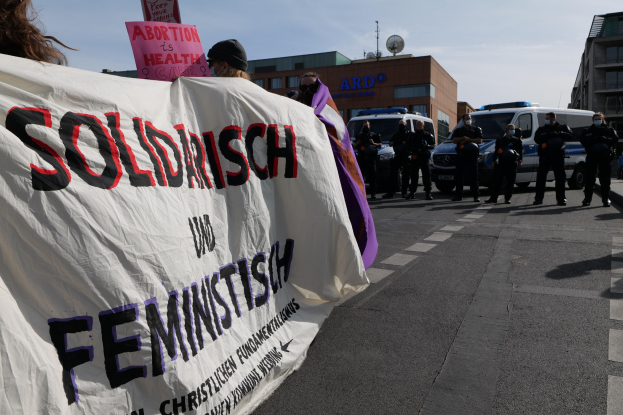 Gruppe von Menschen marschiert die Straße entlang und hält ein "Solidarität und Feminismus"-Schild, mit geparkten Fahrzeugen, Gebäuden, einer Schüsselantenne und einem klaren blauen Himmel im Hintergrund.