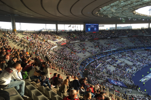 Große Menschenmenge im Allianz Arena Stadion bei einem Fußballspiel sitzend, mit einer Bühne rechts, Flaggen, Stangen und einem Bildschirm im Hintergrund und Himmel oben sichtbar.