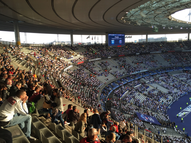 Große Menschenmenge im Allianz Arena Stadion bei einem Fußballspiel sitzend, mit einer Bühne rechts, Flaggen, Stangen und einem Bildschirm im Hintergrund und Himmel oben sichtbar.