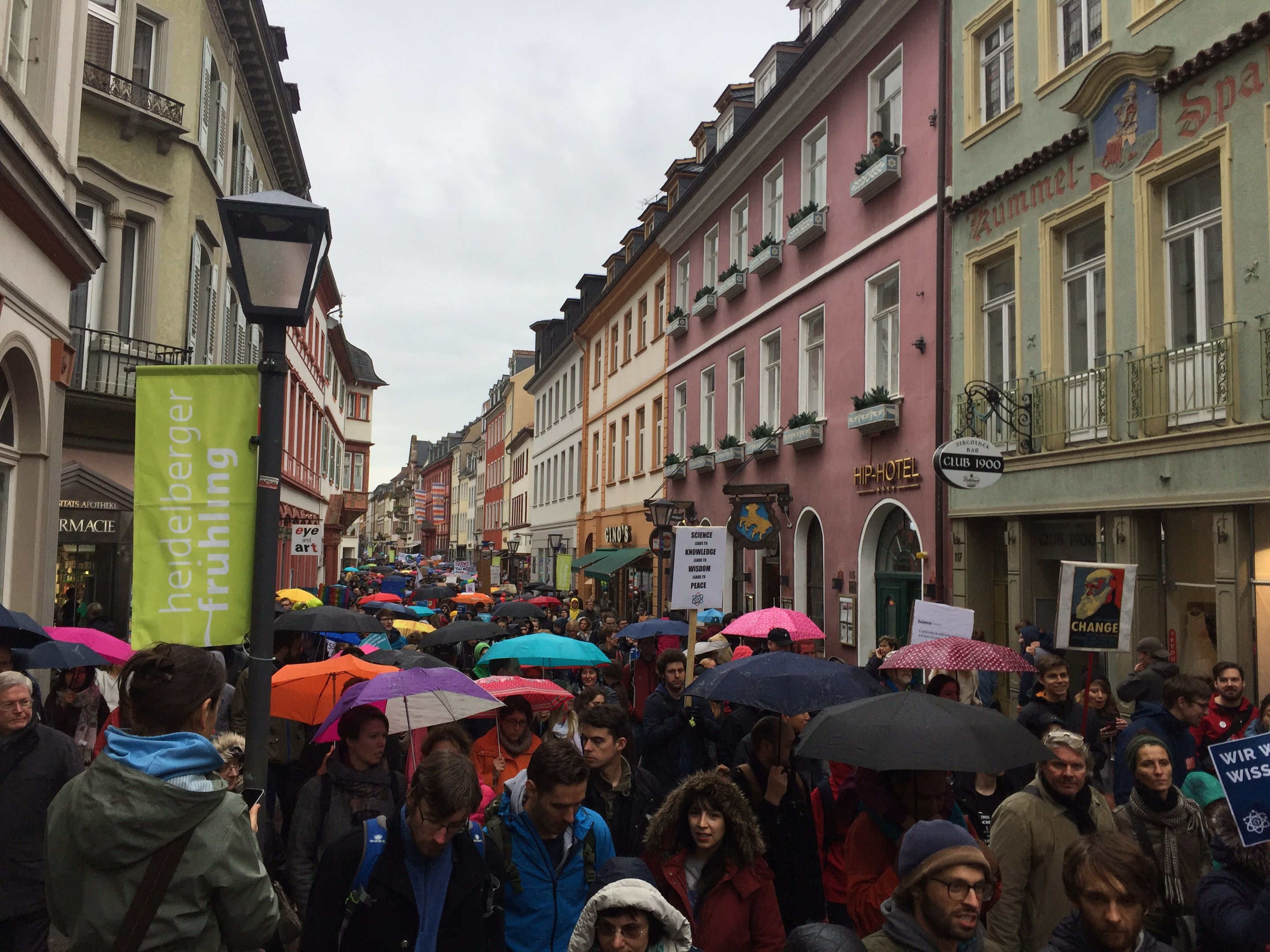 Große Gruppe von Menschen mit Schirmen auf einer Straße unterwegs, einige mit Taschen und Schildern, mit Gebäuden und einem Laternenmast im Hintergrund.