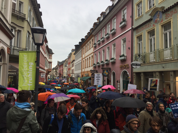 Große Gruppe von Menschen mit Schirmen auf einer Straße unterwegs, einige mit Taschen und Schildern, mit Gebäuden und einem Laternenmast im Hintergrund.