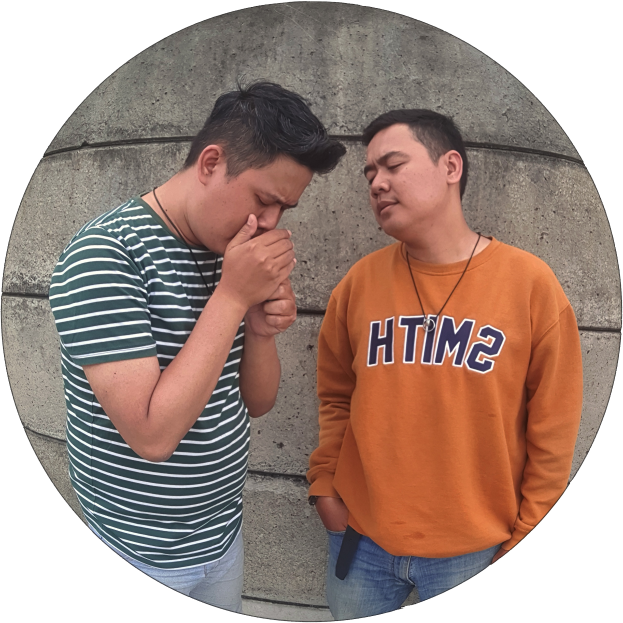 Zwei Männer stehen Seite an Seite vor einer Wand, einer in einem orangen T-Shirt und blauen Jeans, der andere in einem grün-weißen gestreiften Hemd, beide wirkend besorgt und in eine hitzige Diskussion vertieft.