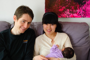 Eine Frau in einem weißen Hemd hält ein Baby und lächelt, waß sie auf einem Sofa sitzt, neben einem Mann in einem schwarzen T-Shirt, der ebenfalls lächelt, mit einem rosafarbenen Fotoраhmen an der weißen Wand dahinter.