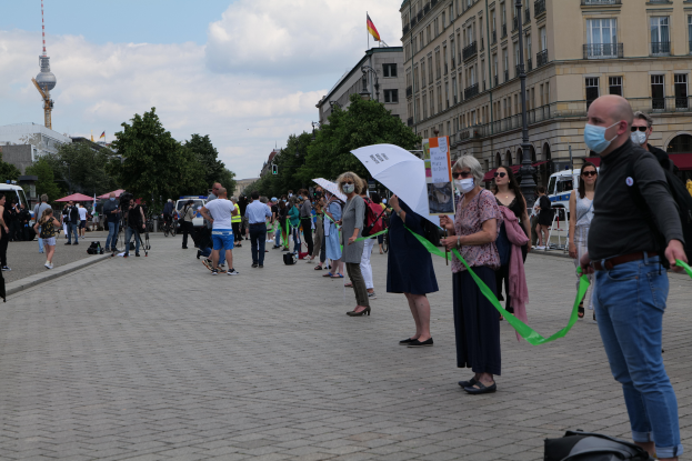 Eine Gruppe von Menschen marschiert auf einer Berliner Straße und hält Schilder und Transparente, einige tragen Masken und Taschen oder Schirme, mit Bäumen, Gebäuden, geparkten Fahrzeugen und einem turmartigen Gebäude mit Fahnen im Hintergrund bei bewölktem Himmel.