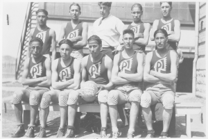 Ein Schwarz-Weiß-Foto einer Basketballmannschaft, die auf einer Bank sitzt, mit einem Mann in der Mitte, der einen Ball hält, Treppen und eine Wand im Hintergrund und Text unten im Bild.