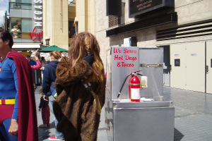 Eine Menschenmenge, einige in Kostümen, versammelte sich vor Gebäuden mit sichtbarer Beschriftung, einer Box mit Schrift und einem Feuerlöscher.