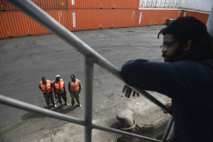 Ein Mann in legerer Kleidung steht auf einem Metallgelönder in der Nähe einer Gruppe von Menschen mit orangen Jacken und Kappen, mit Containern und einer Wand im Hintergrund am Hafen von Los Angeles.