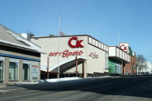 Außenansicht eines Stadt-Sportgeschäfts an einer Straßenecke mit umliegenden Gebäuden, Straßenbefestigungen und schneebedeckter Straße unter einem sichtbaren Himmel.