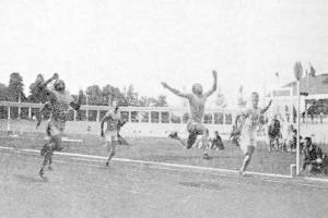 Ein Schwarz-Weiß-Foto von Männern, die auf einem Feld Fußball spielen, mit Zuschauern, Pfosten, Fahnen, Gebäuden, Bäumen und einem klaren Himmel im Hintergrund und dem Text "Sydney Football Club" unten.
