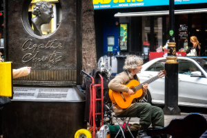 Ein Mann sitzt im Freien und spielt Gitarre, links neben ihm eine Statue, im Hintergrund ein Baum und rechts ein weißes Auto und Geschäfte.