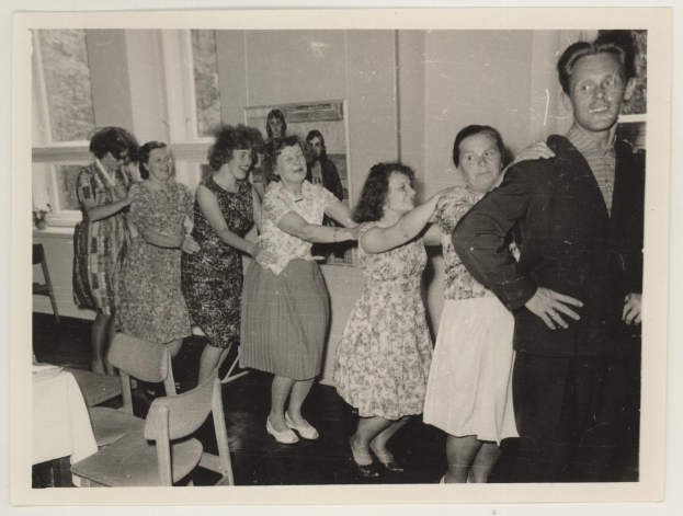 Gruppe von Menschen, die in einem räumlichen Bereich mit Stühlen und einem mit weißer Decke bedeckten Tisch tanzen. Im Hintergrund befindet sich eine Wand mit einem Rahmen und Fenstern.