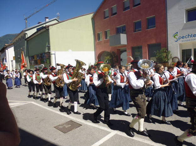 Eine Gruppe von Menschen in traditioneller bayrischer Tracht, die auf einer Straße mit Gebäuden Musikinstrumente spielen, einige halten Fahnen, mit einem Hügel und einem klaren blauen Himmel im Hintergrund.