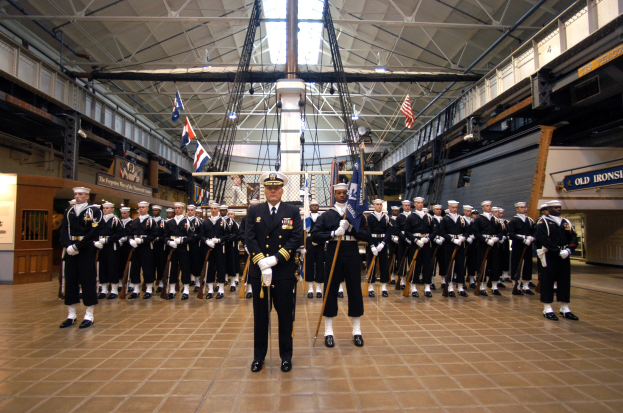 Eine Gruppe uniformierter Männer in einem großen Raum mit Gewehren, Flaggen und Schildern an den Wänden und Deckenleuchten und Stangen.