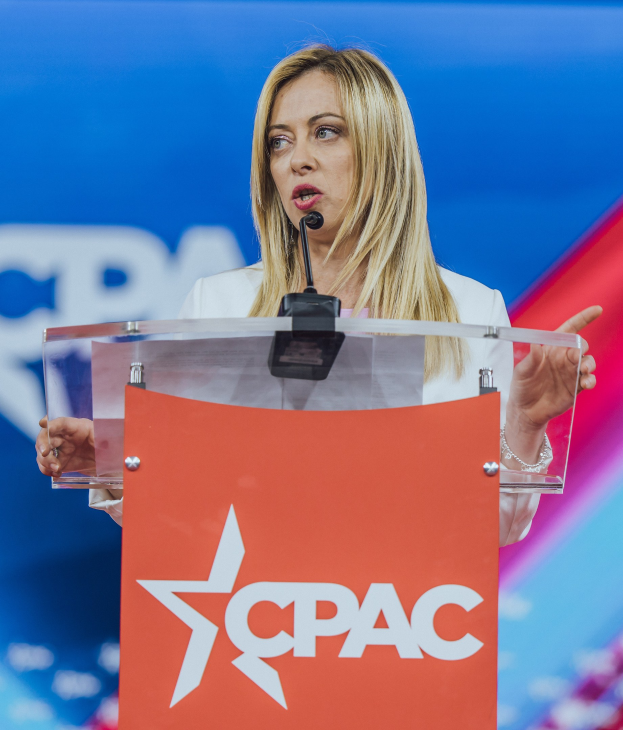 Frau in weißem Kleid, die an einem Rednerpult mit Mikrofon spricht, unscharfer Hintergrund.