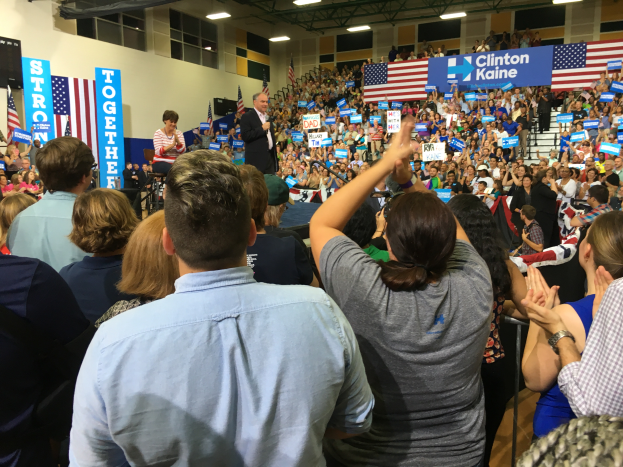Eine große Menschenmenge steht vor einer Bühne, ein Mann spricht in ein Mikrofon und eine Frau steht neben ihm, während Fahnen, Banner und ein Lautsprecherbox auf der Bühne zu sehen sind und die Menge klatscht und jubelt.