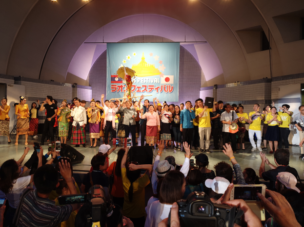 Eine Gruppe von Menschen auf einer Bühne, einige halten Kameras und Telefone, andere Musikinstrumente, mit einem Banner und Lichtern im Hintergrund, was eine Festival-Eröffnung vermuten lässt.