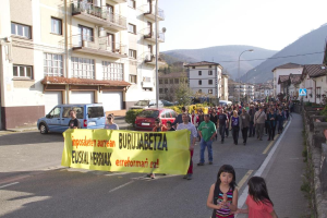 Eine Straße außerhalb der Stadt mit zwei Mädchen unten, Menschen in der Mitte mit einer gelben Fahne, geparkte Fahrzeuge links, Sträucher, Bäume, Mäste, Gebäude, Straßenlaternen und Hügel im Hintergrund, unter einem Himmel oben.