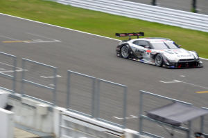 Ein Nissan GT-R Nismo GT3 Rennwagen auf der Nürburgring 24 Stunden Strecke, mit einem Zaun und Gras auf beiden Seiten, einem Schuppen und ein paar Menschen unten und einem unscharfen Hintergrund.