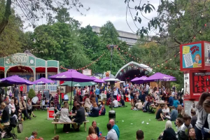 Eine große Gruppe von Menschen sitzt auf einem grünen Feld mit Schirmen, Tischen und einer Tafel, die ein Biergarten in London anzeigt.