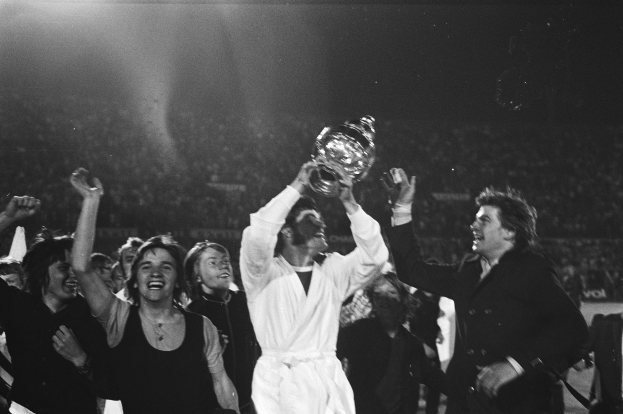 Eine Gruppe von Menschen feiert mit einem Pokal vor einer jubelnden Menge, die zentrale Figur hält ihn triumphierend hoch, während andere jubeln und klatschen, die Szene in Schwarz-Weiß mit heller Beleuchtung.