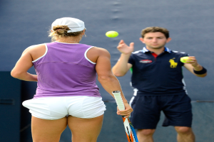 Eine Frau in einem lila T-Shirt, weißen Shorts und einer Mütze hält einen Schläger in der linken Hand, während ein Mann in einem blauen Jersey einen Ball in ihre Richtung wirft.