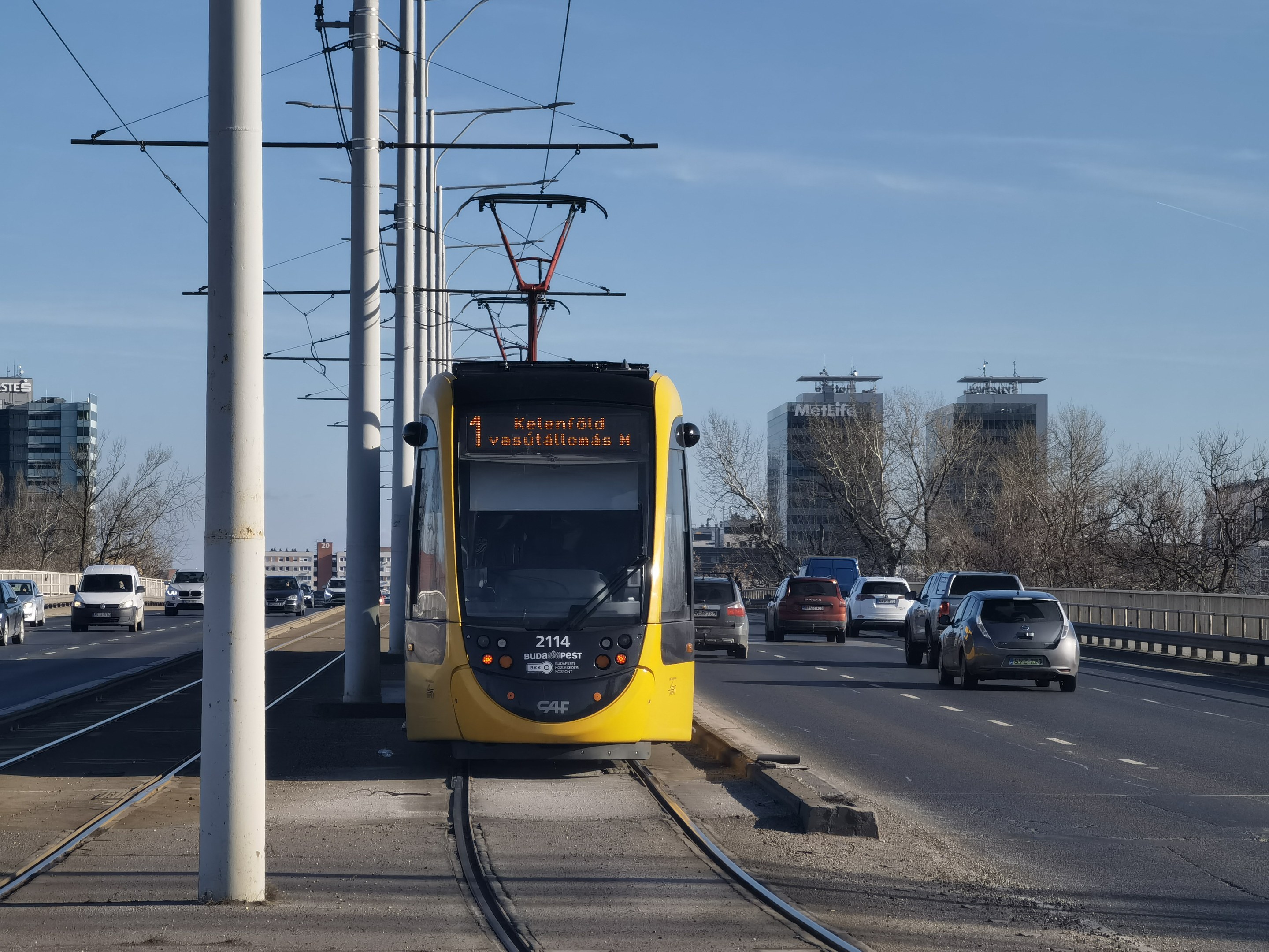 Gelber Straßenbahnwagen auf einer Stadtstraße mit Autos, Strommasten, Bäumen, Gebäuden und einem klaren blauen Himmel im Hintergrund.