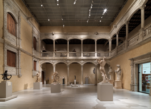 Großer Raum in einem Museum mit zahlreichen Statuen und Skulpturen auf Sockeln, architektonischen Elementen wie Wänden, Säulen und Geländern, Deckenleuchten und einigen Besuchern.