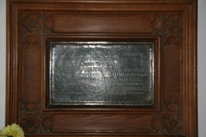 Eine Gedenktafel mit Text ist an einer braunen Wand oder Holztür angebracht, mit einem gelben Objekt, das einer Blumenvase ähnelt, in der linken unteren Ecke.