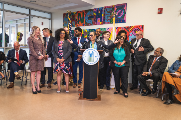Eine Gruppe von Menschen, die vor einem Podium mit Mikrofon und einer Tafel mit der Aufschrift "Sancho-Stadtrat tritt dem Stadtrat bei" stehen, mit Flaggen, Feuerlöscher und Deckenleuchten an der Rückwand.