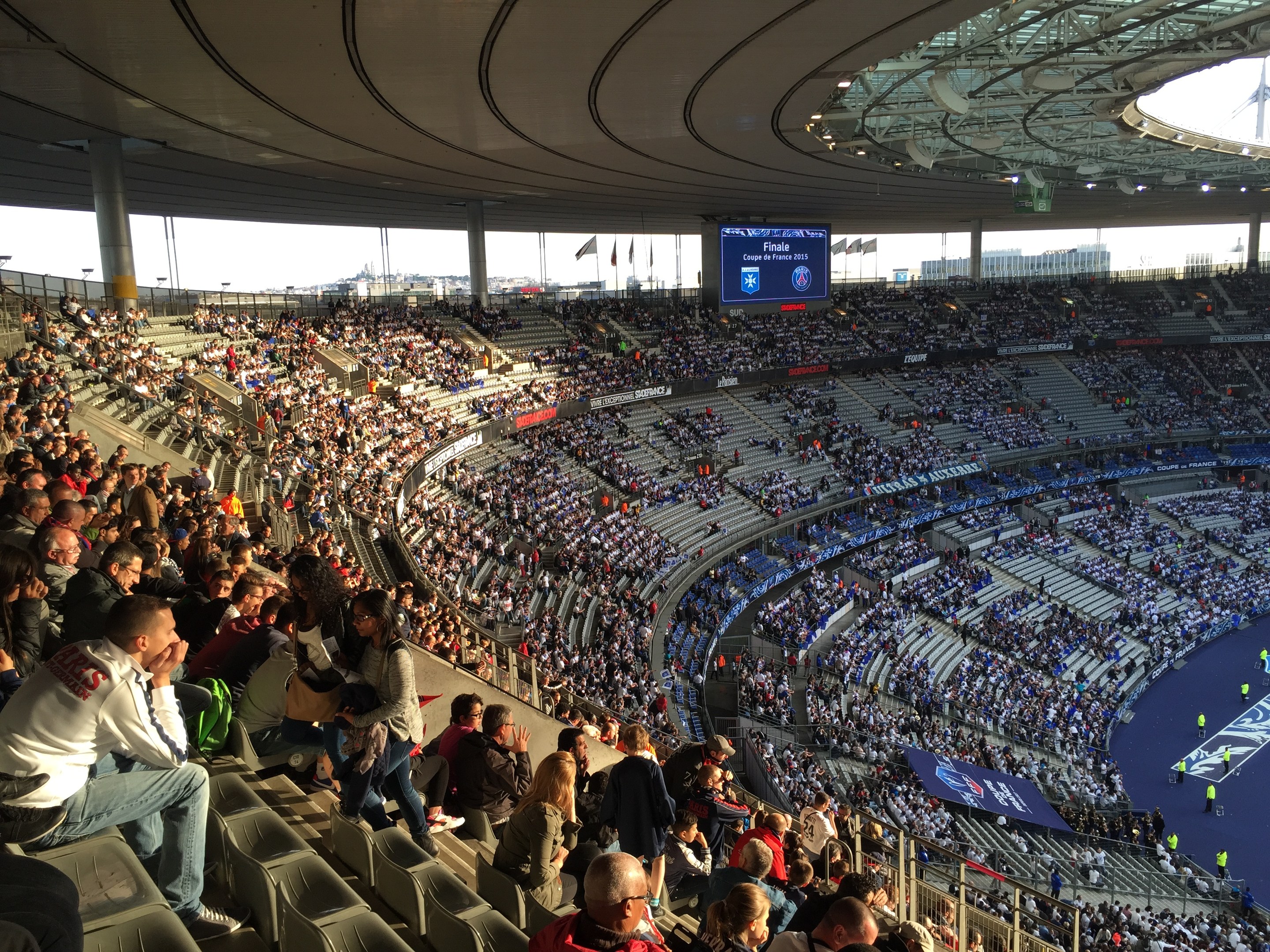 Große Menge im Allianz Arena Stadion bei einem Fussballspiel sitzend, mit einer Bühne und Fahnen auf der rechten Seite, einem Bildschirm im Hintergrund und Himmel über ihnen.