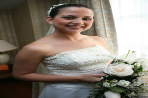 Eine Braut steht lachend mit einem Strauß in der Hand vor einem Vorhang und einer Wand und einer Lampe auf einem Tisch links.