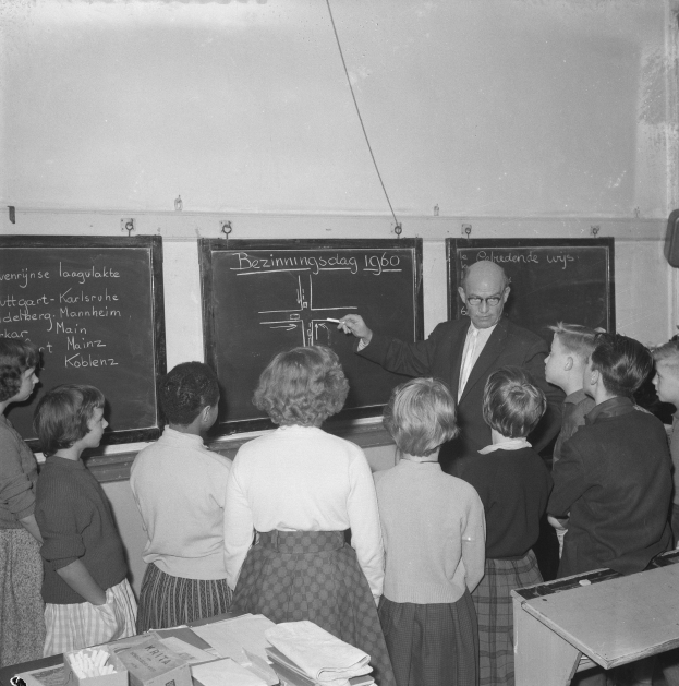Schwarzes und weißes Foto eines Lehrers, der vor einer Tafel steht und einen Marker hält, während Kinder an einem Tisch mit Büchern und Materialien sitzen; ein an der Wand befestigter Lautsprecher ist auf der rechten Seite zu sehen.