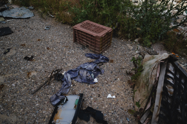 Haufen Müll einschließlich einer Plastikbox, Kleidung und anderen Gegenständen neben einer Straße neben einem Zaun, mit Pflanzen und Gras im Hintergrund.