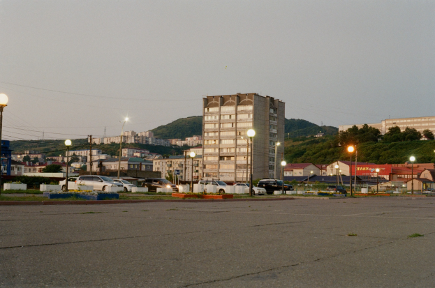Leerer Parkplatz mit Autos, umgeben von Gebäuden, Straßenlaternen, Strommasten, Stromkabeln, Bäumen, Hügeln und einem klaren blauen Himmel.