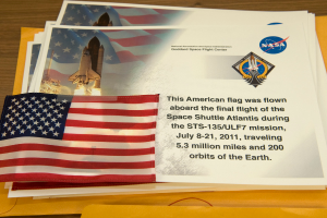 Ein zentrales Element sind Plakate mit Text, einer Flagge, einem Raketenmodell und verschiedenen Logos, mit einem Tisch unten und einem Boden oben im Bild.