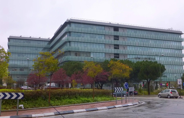Großes modernes Gebäude mit zahlreichen Fenstern, identifiziert als Hauptquartier der Europäischen Zentralbank, umgeben von Grünflächen, Straßeninfrastruktur und Fahrzeugen unter einem bewölkten Himmel.