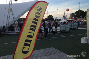 Ein buntes Volksfest-Szenario mit Zelten, einem zentralen Mast, Menschen, Bäumen, Strommasten, Straßenbahnen auf der Straße und einem Eisenständer auf der linken Seite unter einem klaren Himmel.