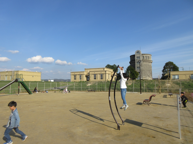 Eine Gruppe von Kindern, die auf einem Spielplatz vor einem Gebäude mit Fenstern spielt, umgeben von einem Metallzaun, Bänken, einer Treppe, einem Metallpfahl, einigen Pflanzen mit Blumen, Gras, der Rinde eines Baumes, einigen Pfählen, einem Turm, einigen Bäumen und einem bewölkten Himmel.