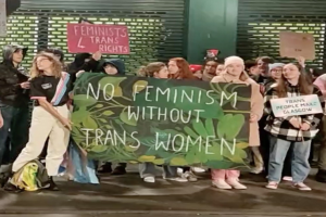 Eine Gruppe von Menschen hält ein Banner mit der Aufschrift "Kein Feminismus ohne Trans-Frauen" und verschiedene Schilder in einer öffentlichen Fläche.
