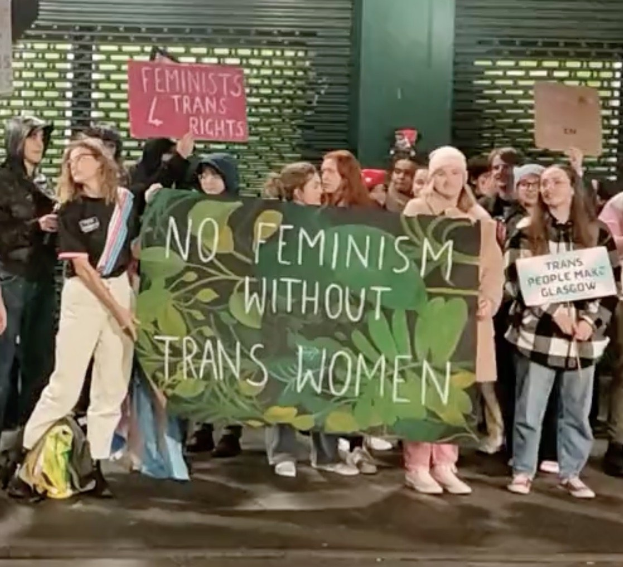 Eine Gruppe von Menschen hält ein Banner mit der Aufschrift "Kein Feminismus ohne Trans-Frauen" und verschiedene Schilder in einer öffentlichen Fläche.