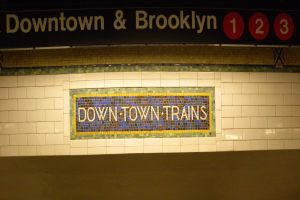 Eine U-Bahn-Station mit einem "Downtown & Brooklyn"-Schild an der Wand und einer Tafel mit Text und Zahlen oben.