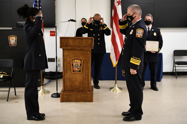 Gruppe uniformierter Personen in Masken vor einem Podium mit einem Mikrofon, umgeben von Flaggen und Möbeln, während einer Vereidigung, bei der eine Person ein Zertifikat hält.