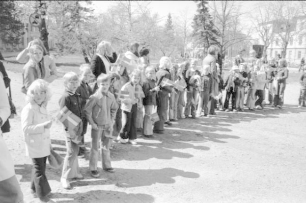 Schwarz-weißes Bild einer Protestdemo auf einem Schulgelände, mit Menschen, die Fahnen in einer Reihe auf einem Schotterweg halten, Bäume und Gebäude im Hintergrund unter einem klaren Himmel.