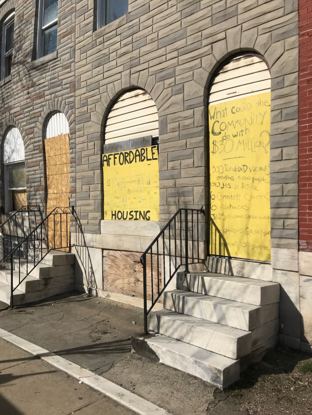 Verschlossene Fenster an einem Gebäude mit einer Tafel, auf der 'Bezahlbarer Wohnraum' steht, Treppe mit Geländer, die zum Eingang führt, und Straße unten.