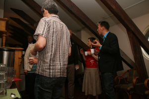 Mehrere Personen mit Tassen, links ein Becher und ein Tisch, oben ein Dach und rechts Stühle.