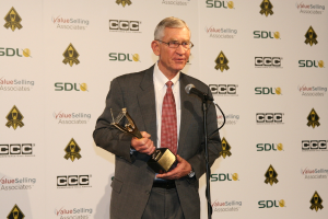 Ein Mann mit Brille steht vor einem Mikrofon und hält eine Trophäe in der Hand, mit einem Plakat im Hintergrund.