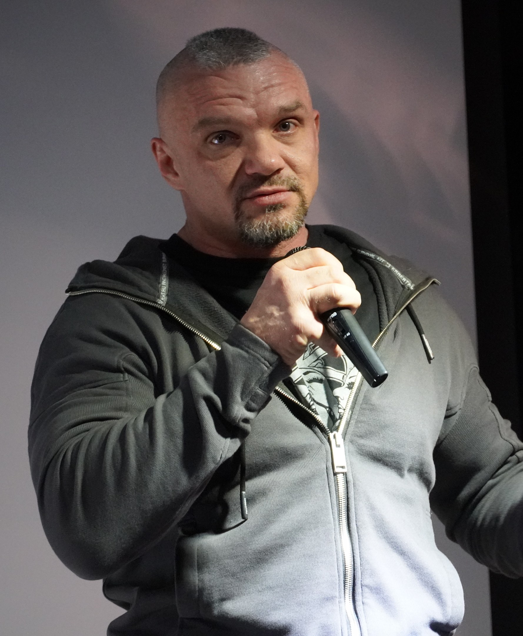Ein Mann in einer schwarzen Kapuzenjacke und einem schwarzen Anzug mit einem weißen Hemd, der ein Mikrofon hält, steht selbstbewusst vor einer Wand und sieht aus wie ein Komiker.