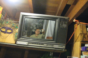 Ein Fernsehbildschirm zeigt eine Person umgeben von Pfauenfedern, Büchern und Kartons.